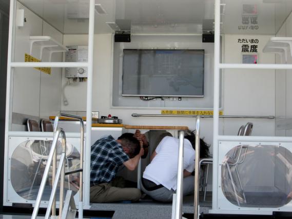 起震車で地震の揺れを体験し、テーブルの下にもぐっている人々の写真