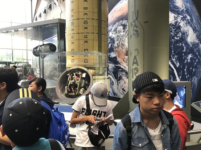 博物館内を見学する訪問団と壁の大きな地球の写真やロケットなどの展示物の写真